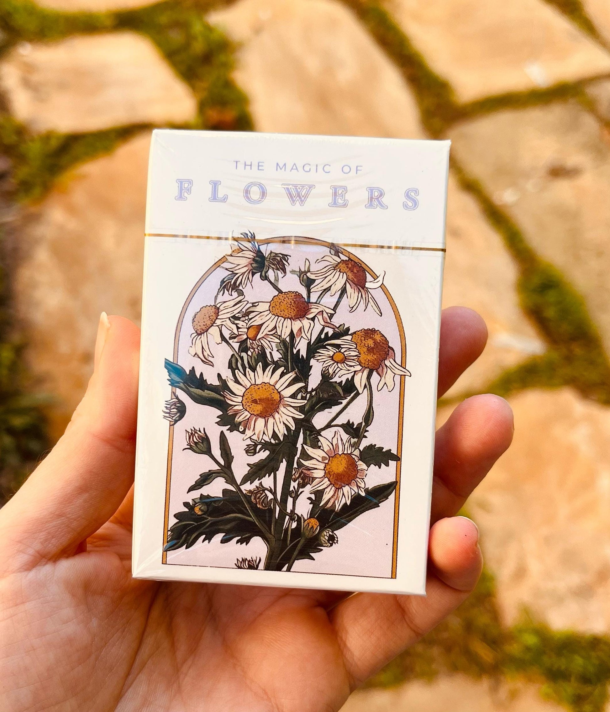 A deck of cards with a floral design. The box is white with gold trim and features an illustration of daisies in a gold-framed arch. The text "THE MAGIC OF FLOWERS" is printed at the top in lavender letters.