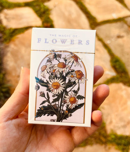 A deck of cards with a floral design. The box is white with gold trim and features an illustration of daisies in a gold-framed arch. The text "THE MAGIC OF FLOWERS" is printed at the top in lavender letters.
