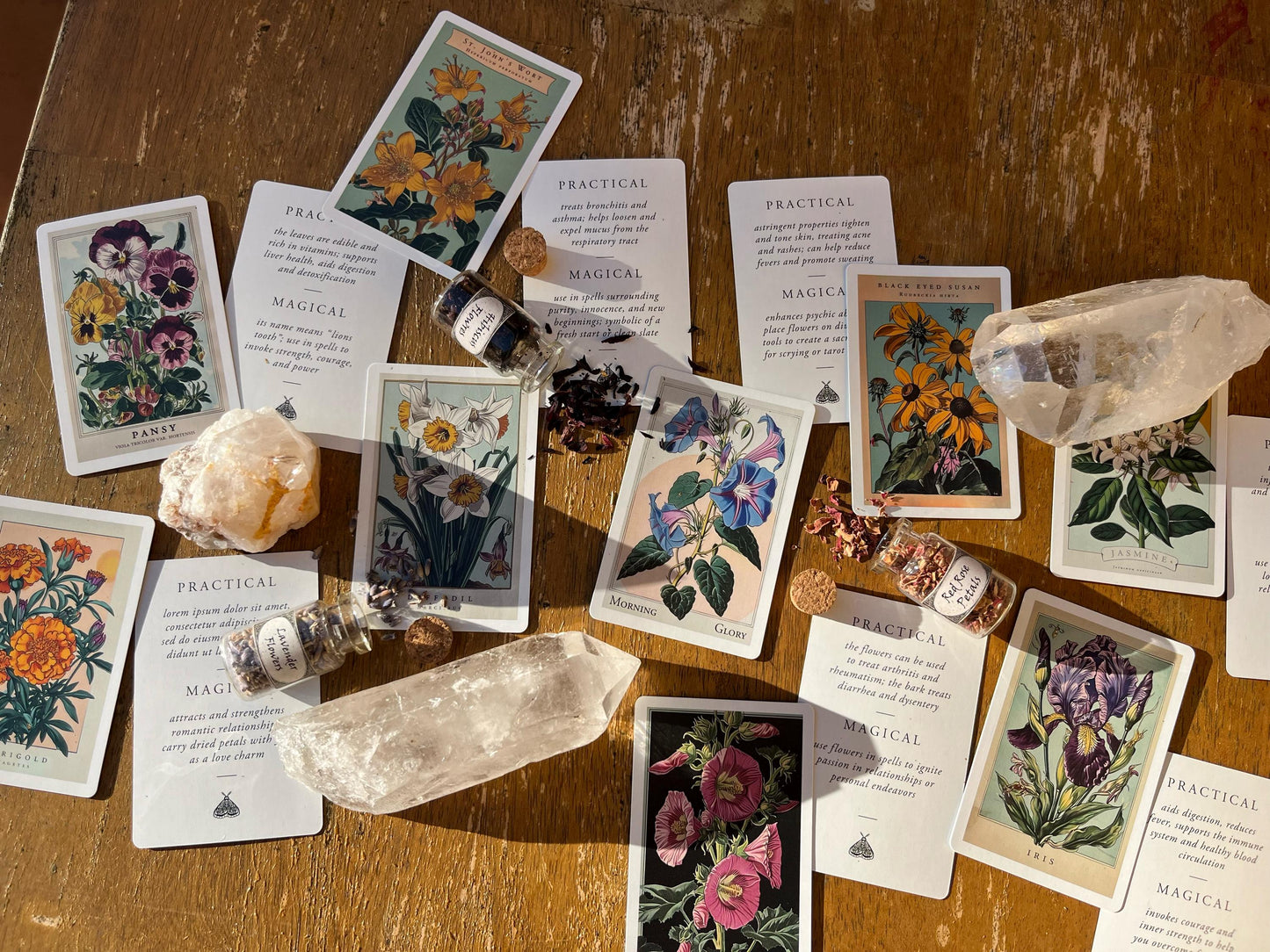 A collection of botanical-themed oracle cards, crystals, and small glass bottles with cork stoppers, arranged on a wooden surface. The cards feature floral illustrations and text. The bottles contain dried petals.