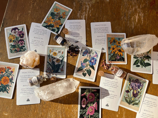 A collection of botanical-themed oracle cards, crystals, and small glass bottles with cork stoppers, arranged on a wooden surface. The cards feature floral illustrations and text. The bottles contain dried petals.